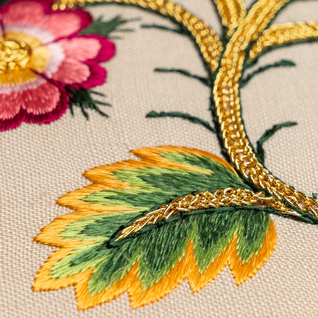 Close-up of embroidered floral design with pink flower, green leaves, and gold thread on beige fabric.