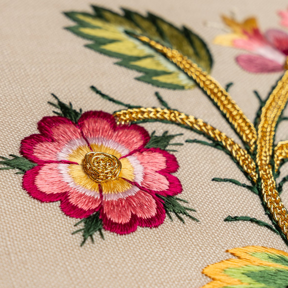 Close-up of detailed floral embroidery with pink flower and gold leaves on beige fabric.